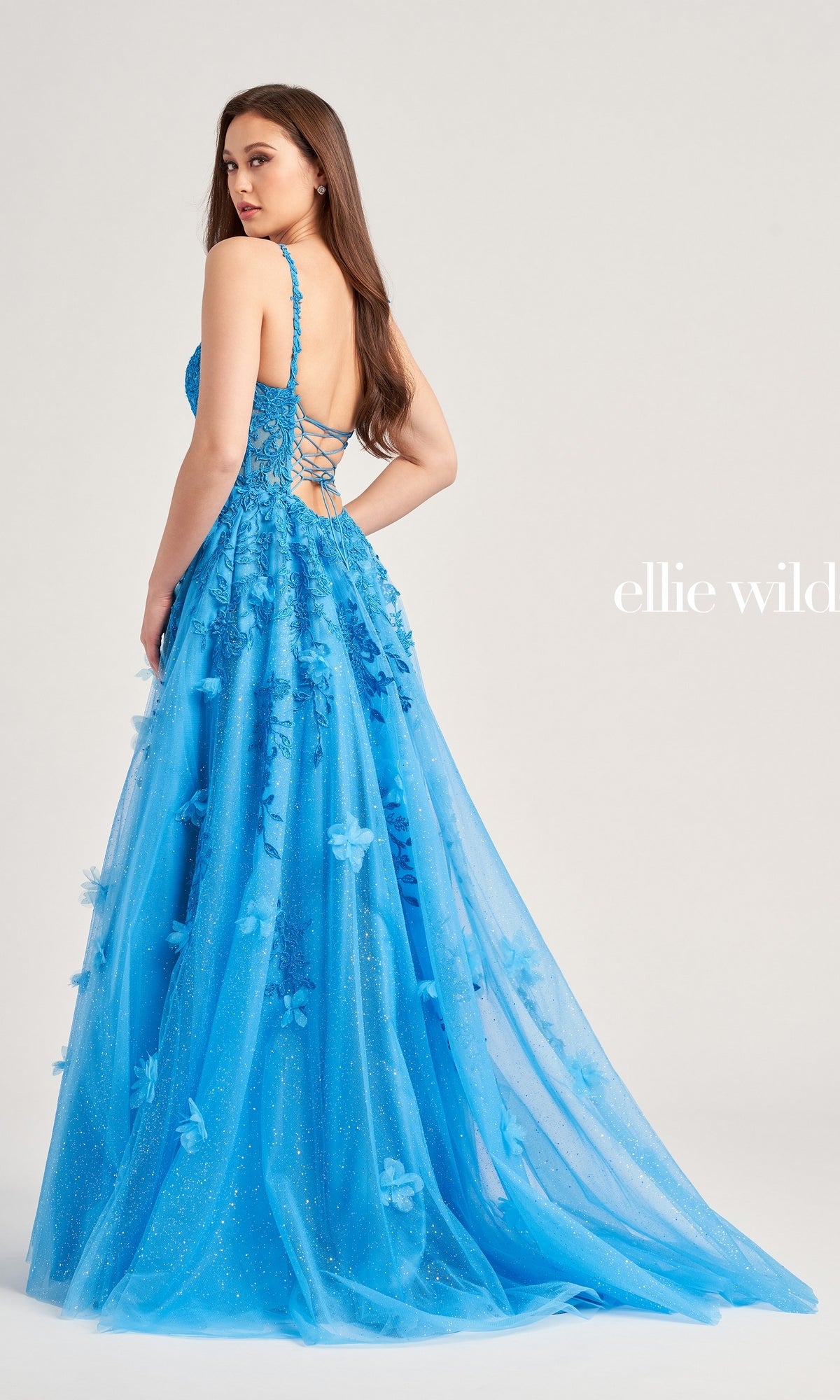 A woman stands against a plain background wearing the Glitter-Tulle Ellie Wilde Long Prom Gown EW35081, featuring bright blue glitter tulle, floral appliqué, a lace-up back, and a tulle overlay.